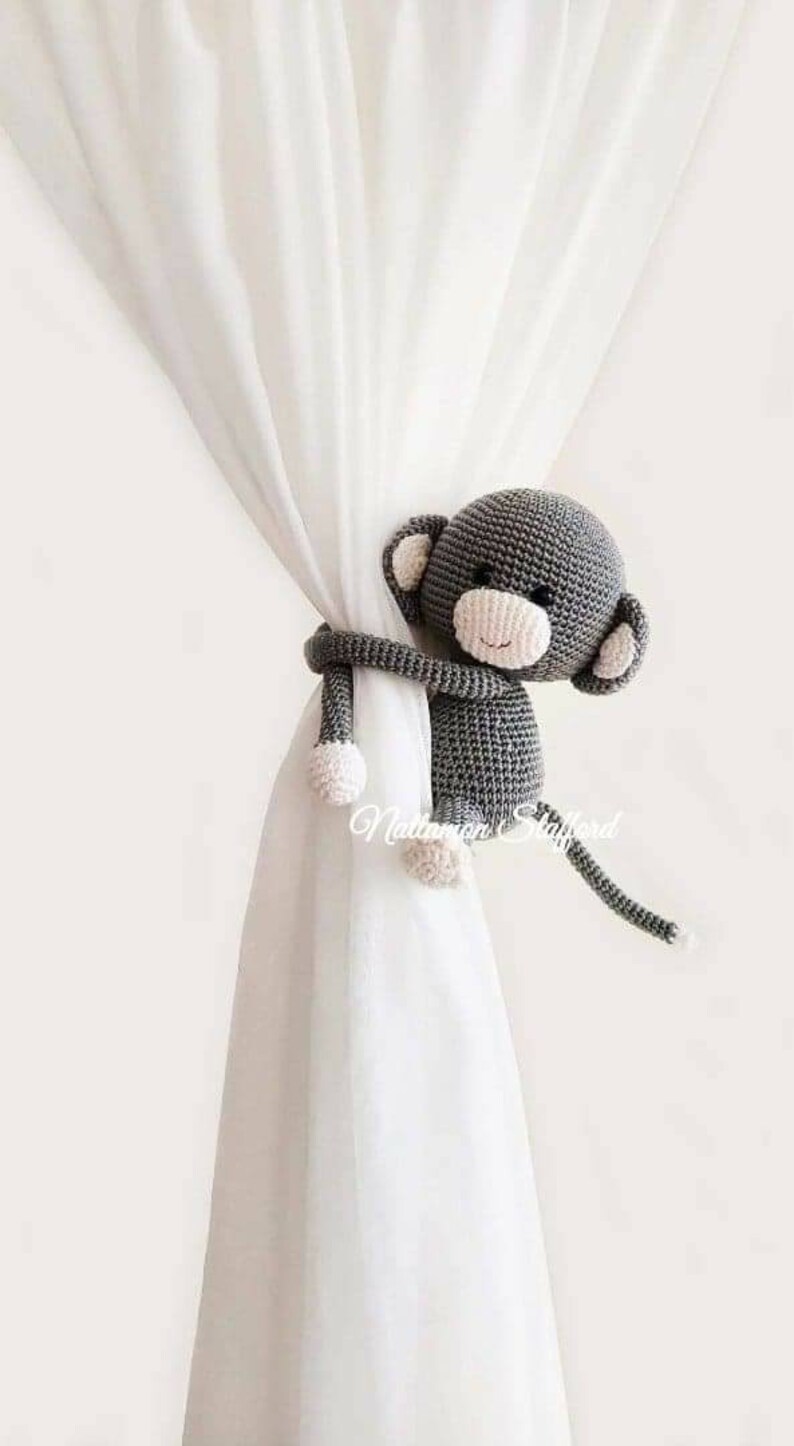 May include: A gray crocheted monkey toy with pink accents is used as a curtain tieback. The monkey is holding the curtain with its arms and has a long tail.