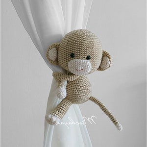 May include: A crocheted monkey toy in beige with white accents is used as a curtain tieback. The monkey is holding onto the curtain with one arm and its tail is draped over the curtain.