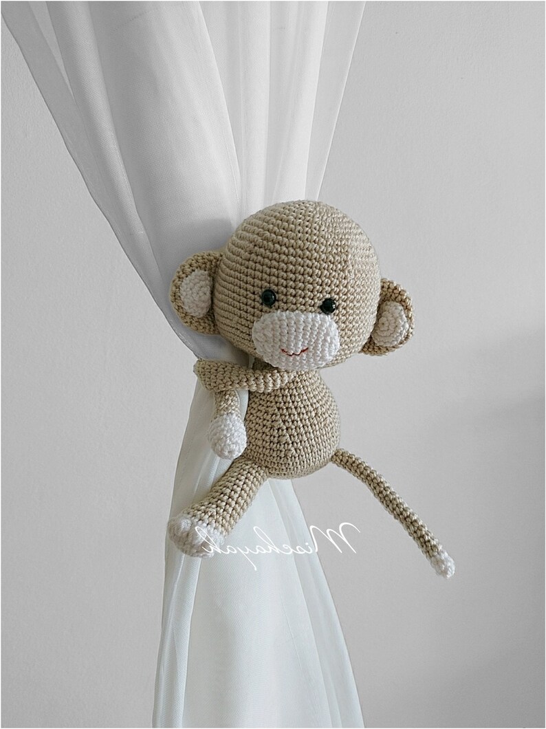 May include: A crocheted monkey toy in beige with white accents is used as a curtain tieback. The monkey is holding onto the curtain with one arm and its tail is draped over the curtain.