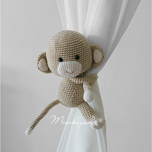 May include: A crocheted monkey toy in beige and white, holding a curtain with its arms. The monkey has a long tail and is used as a curtain tieback.