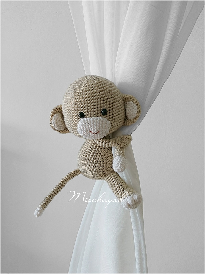 May include: A crocheted monkey toy in beige and white, holding a curtain with its arms. The monkey has a long tail and is used as a curtain tieback.