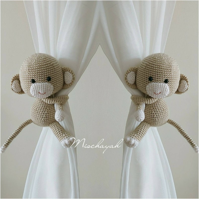 May include: Two crocheted monkey curtain tiebacks in beige with white accents. The monkeys are holding onto the white curtains with their arms and have their tails hanging down.