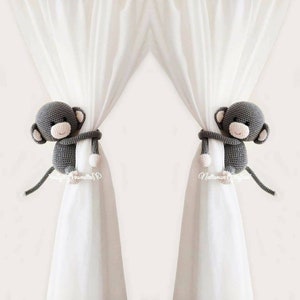 May include: Two gray crocheted monkey curtain tiebacks with white yarn accents. The monkeys are holding onto white curtains.