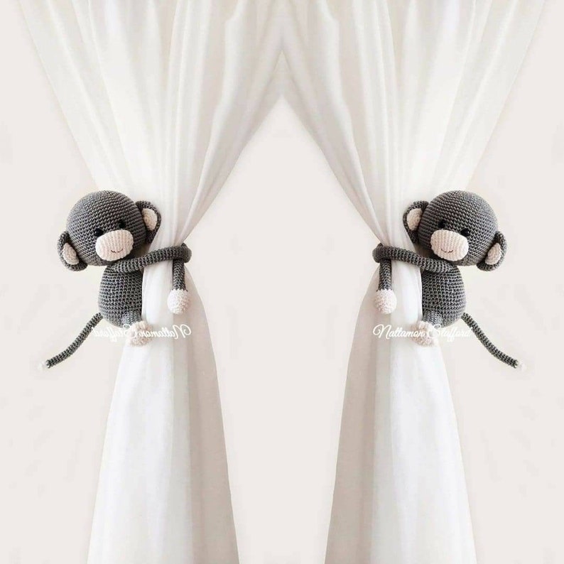 May include: Two gray crocheted monkey curtain tiebacks with white yarn accents. The monkeys are holding onto white curtains.