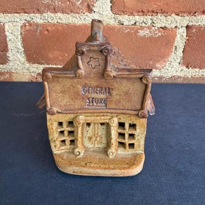 May include: A ceramic miniature of a general store, featuring a tan and brown color scheme. The building has a sign that reads "GENERAL STORE" and a textured roof. The front of the store has a door and window cutouts.