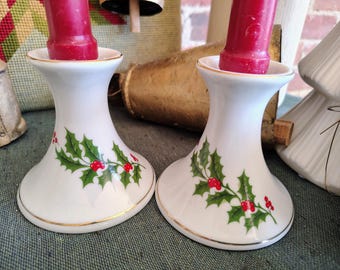 May include: Two white ceramic candle holders with a gold rim and a holly berry design. Each holder has a red candle inserted. The holders are on a green surface.