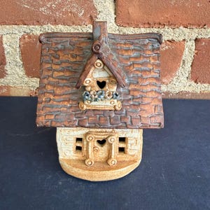 May include: A ceramic house figurine with a brown, textured roof and a tan base. The house features a heart-shaped window and a small birdhouse with blue floral accents. The figurine is set against a brick background.
