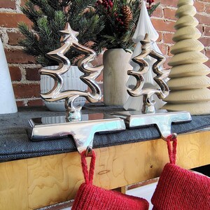 May include: Two silver Christmas tree-shaped stocking holders with red stockings hanging from them. The holders are on a wooden surface with decorative trees and greenery in the background. The holders are made of shiny metal.