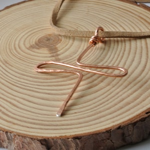May include: A copper pendant necklace with an abstract cross design. The pendant hangs from a brown leather cord, displayed on a natural wood slice. The copper has a hammered texture and a bright, metallic finish. The necklace is a unique piece of jewellery.