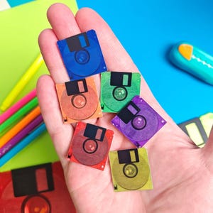 May include: A hand holding several miniature, colorful floppy disks. The disks are in various colors including blue, orange, green, purple, red, and yellow. The disks have a black slider and a circular design in the center.