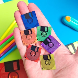 May include: A hand holding several miniature, colorful floppy disks. The disks are in various colors including blue, orange, green, purple, red, and yellow. The disks have a black slider and a circular design in the center.