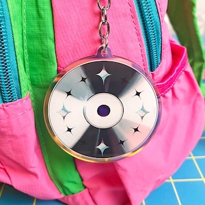 May include: A circular acrylic keychain featuring a graphic of a silver compact disc with a dark center and black four-pointed star accents. The keychain is attached to a silver metal split ring and chain, hanging from a bright pink and green backpack.