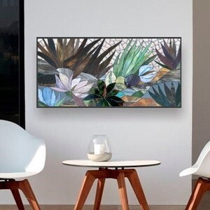May include: A white mosaic art piece depicting a tropical scene with green and brown leaves and blue flowers. The art piece is hanging on a white wall above a small wooden table and two white chairs.