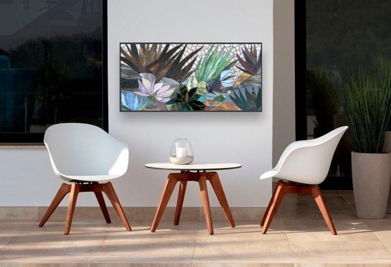 May include: A white mosaic art piece depicting a tropical scene with green and brown leaves and blue flowers. The art piece is hanging on a white wall above a small wooden table and two white chairs.