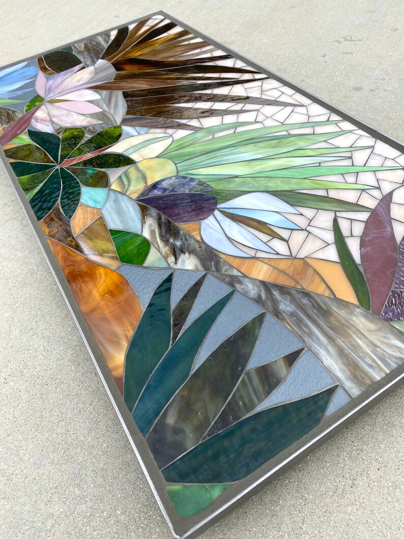 May include: A rectangular mosaic artwork featuring a variety of colors and textures, including green, brown, purple, and white. The mosaic depicts a stylized floral design with leaves and flowers.