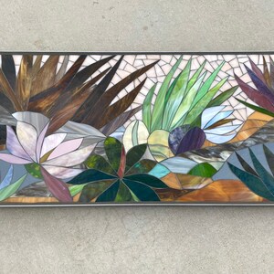 May include: A rectangular mosaic artwork featuring a variety of plants and flowers. The artwork is made of small pieces of glass in shades of brown, green, blue, purple, and white. The artwork is framed in a silver metal frame.