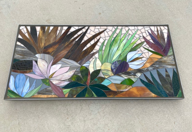 May include: A rectangular mosaic artwork featuring a variety of plants and flowers. The artwork is made of small pieces of glass in shades of brown, green, blue, purple, and white. The artwork is framed in a silver metal frame.
