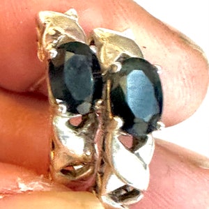 May include: A pair of silver-toned earrings featuring oval-cut, dark black stones set in a metallic frame. The earrings have a textured, braided-style metal band design that supports the central dark gemstone setting on each piece.