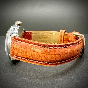 Puede incluir: Un reloj de pulsera vintage con caja plateada y correa de cuero naranja texturizada. La correa est&aacute; cosida con hilo en contraste y tiene una hebilla plateada. El reloj se encuentra sobre un fondo oscuro.