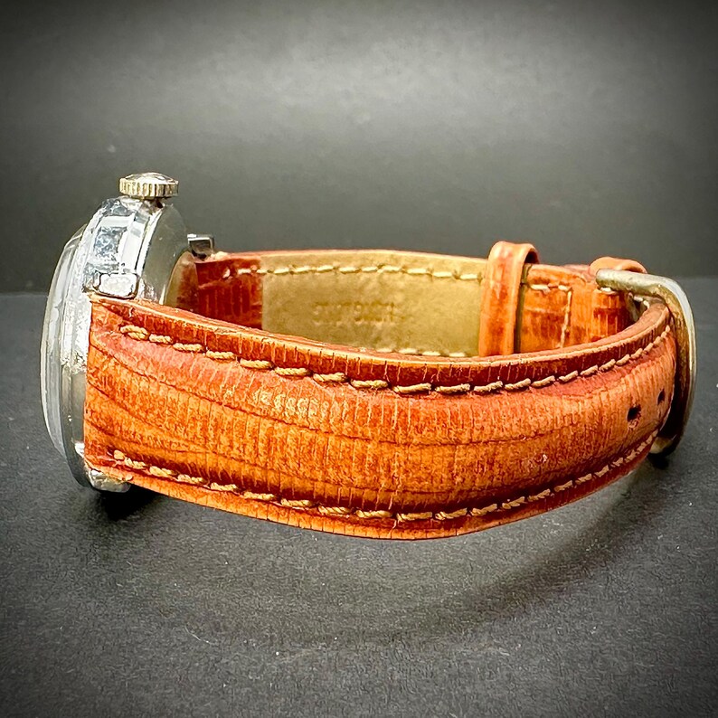 Puede incluir: Un reloj de pulsera vintage con caja plateada y correa de cuero naranja texturizada. La correa est&aacute; cosida con hilo en contraste y tiene una hebilla plateada. El reloj se encuentra sobre un fondo oscuro.