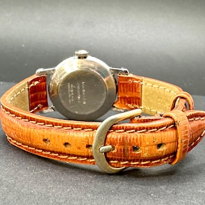 Puede incluir: Un reloj de pulsera vintage con caja plateada y correa de cuero marr&oacute;n anaranjado texturizada. La correa se asegura con una hebilla plateada. La esfera del reloj no es visible en esta imagen.