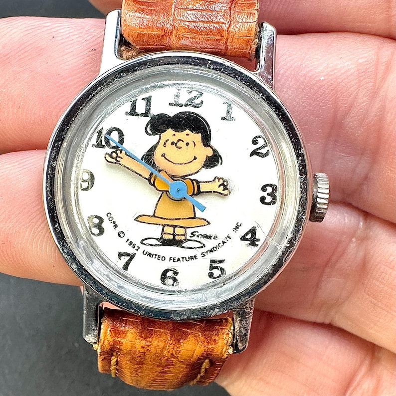 Puede incluir: Reloj de pulsera vintage con un personaje de dibujos animados de Lucy en la esfera. El reloj tiene una esfera blanca con n&uacute;meros negros y una correa de cuero marr&oacute;n. La esfera del reloj incluye el texto "COPR. &copy; 1952 UNITED FEATURE SYNDICATE INC."