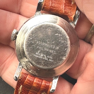 Puede incluir: Primer plano de la parte trasera de un reloj de pulsera vintage con caja de metal plateado y correa de cuero marr&oacute;n. La esfera del reloj est&aacute; inscrita con "ASSEMBLED IN PHILIPPINES", "BASE METAL BEZEL" y "STAINLESS STEEL BACK".