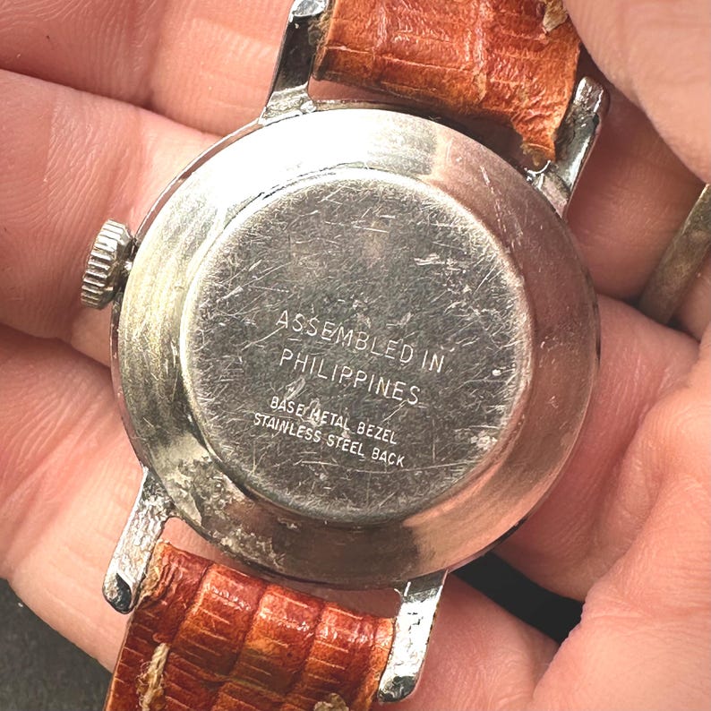 Puede incluir: Primer plano de la parte trasera de un reloj de pulsera vintage con caja de metal plateado y correa de cuero marr&oacute;n. La esfera del reloj est&aacute; inscrita con "ASSEMBLED IN PHILIPPINES", "BASE METAL BEZEL" y "STAINLESS STEEL BACK".