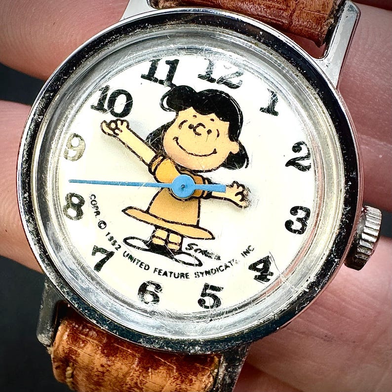 Puede incluir: Reloj de pulsera vintage con un personaje de dibujos animados en la esfera blanca y n&uacute;meros negros. El reloj tiene un bisel plateado y una correa de cuero marr&oacute;n. La esfera del reloj incluye el texto "COPR. &copy; 1952 UNITED FEATURE SYNDICATE, INC."