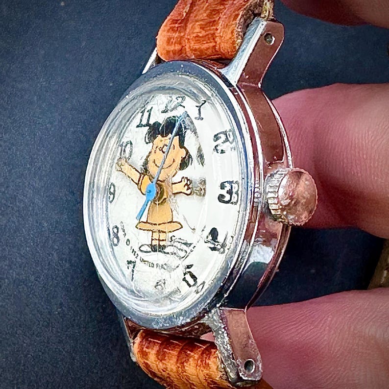 Puede incluir: Reloj de pulsera vintage con caja plateada y correa de cuero marr&oacute;n. La esfera del reloj presenta un personaje de dibujos animados con un vestido amarillo y un segundero azul. El reloj est&aacute; marcado "1952 United Feature Syndicate".