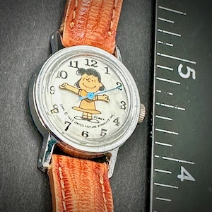 Puede incluir: Un reloj de pulsera vintage con caja plateada y correa de cuero marr&oacute;n. La esfera del reloj presenta una imagen de dibujos animados de Lucy de Peanuts. El reloj tiene una esfera de color claro con n&uacute;meros y agujas negras. La correa mide aproximadamente 1,3 cm de ancho.