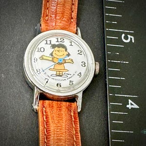 Puede incluir: Reloj de pulsera vintage con caja plateada y correa de cuero marr&oacute;n. La esfera del reloj presenta un personaje de dibujos animados con un vestido amarillo. La esfera es blanca con n&uacute;meros y agujas negras.