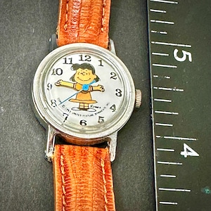 Puede incluir: Un reloj de pulsera vintage con caja plateada y correa de cuero marr&oacute;n. La esfera del reloj presenta un personaje de dibujos animados con un vestido amarillo. La pantalla es anal&oacute;gica cl&aacute;sica con n&uacute;meros negros.