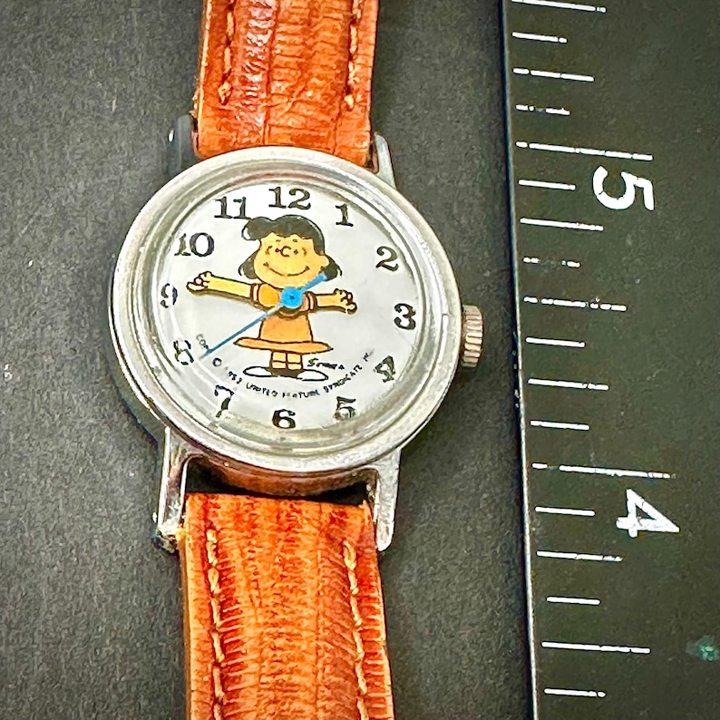 Puede incluir: Un reloj de pulsera vintage con caja plateada y correa de cuero marr&oacute;n. La esfera del reloj presenta un personaje de dibujos animados con un vestido amarillo. La pantalla es anal&oacute;gica cl&aacute;sica con n&uacute;meros negros.
