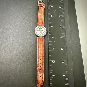 Puede incluir: Un reloj de pulsera plateado con una correa de cuero marr&oacute;n. La esfera del reloj presenta un personaje de dibujos animados. La correa es texturizada y tiene una hebilla. El reloj se muestra sobre una superficie oscura junto a una regla.