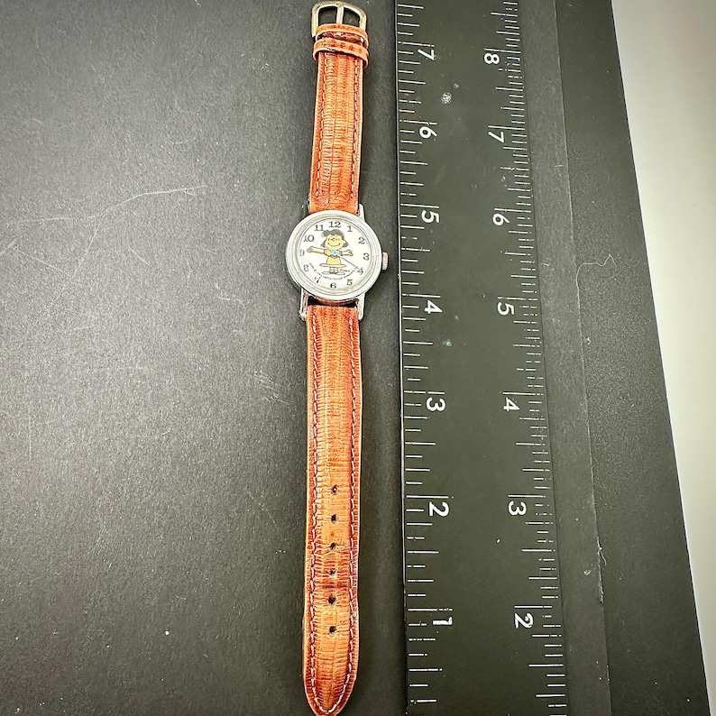Puede incluir: Un reloj de pulsera plateado con una correa de cuero marr&oacute;n. La esfera del reloj presenta un personaje de dibujos animados. La correa es texturizada y tiene una hebilla. El reloj se muestra sobre una superficie oscura junto a una regla.