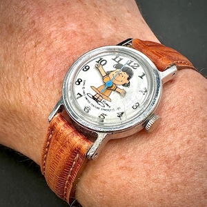 Puede incluir: Reloj de pulsera vintage con caja plateada y correa de cuero marr&oacute;n. La esfera del reloj presenta una ilustraci&oacute;n de un personaje de dibujos animados y n&uacute;meros negros. La correa mide aproximadamente 2 cm de ancho.