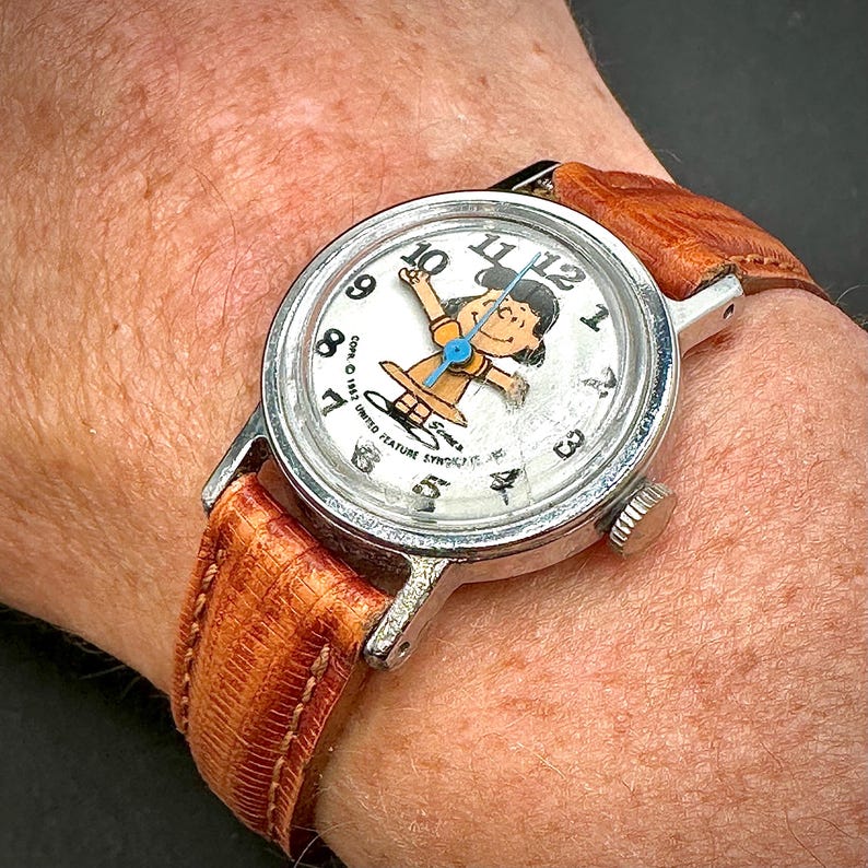 Puede incluir: Reloj de pulsera vintage con caja plateada y correa de cuero marr&oacute;n. La esfera del reloj presenta una ilustraci&oacute;n de un personaje de dibujos animados y n&uacute;meros negros. La correa mide aproximadamente 2 cm de ancho.