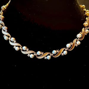 Può includere: Una collana color oro con un motivo ripetuto di perle e piccole gemme trasparenti. La collana ha un design ondulato con perle incastonate tra le sezioni dorate ondulate. Lo sfondo è nero.