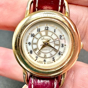 Puede incluir: Reloj de pulsera dorado con esfera blanca y marcadores de hora en números romanos negros. El reloj tiene una correa de cuero roja y dice "Windsor Quartz" en la esfera.