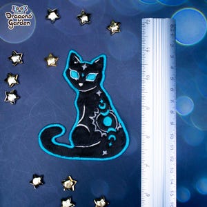 May include: A black cat-shaped patch with a teal outline and celestial designs. The patch is approximately 12 cm tall, with a ruler in the background. The patch is surrounded by star-shaped embellishments and the text "Dragons Garden".