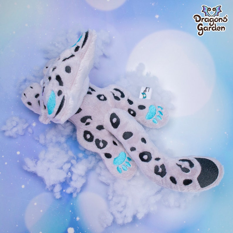 May include: A plush toy in the shape of a snow leopard with grey and black spots. The toy has blue paw pads and a blue spot on its face. The toy is lying on a white fluffy surface.