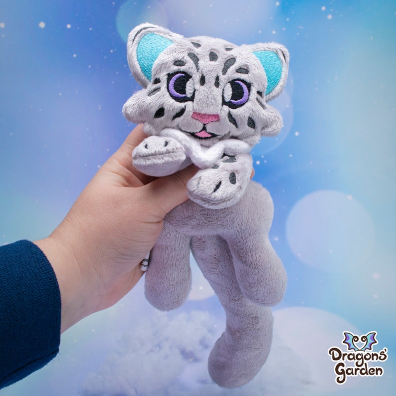 May include: A plush toy of a snow leopard with blue ears and purple eyes. The toy is grey and white with a white belly and a white collar. The toy is being held in a hand.
