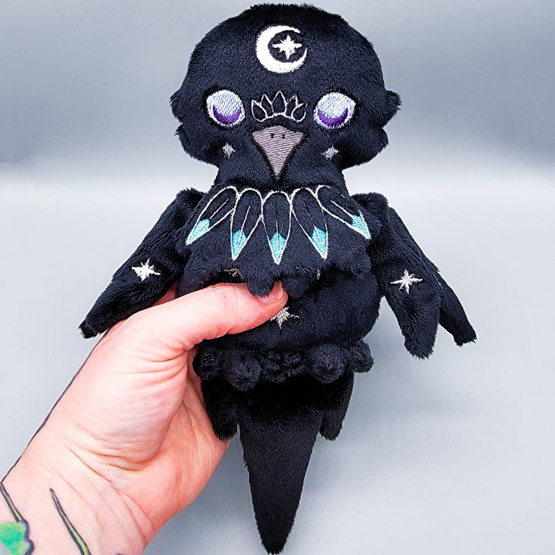 May include: Black plush crow with embroidered details, including a crescent moon, stars, and feathers. The crow has large, purple eyes and a small, gray beak.
