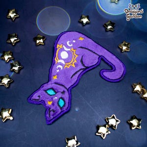 May include: A purple cat-shaped patch with a witch hat design. The hat features a sun, moon, and star motif in yellow and white. The cat's eyes are turquoise. The patch is surrounded by gold star-shaped embellishments. The text "Dragons Garden" is in the upper right corner.