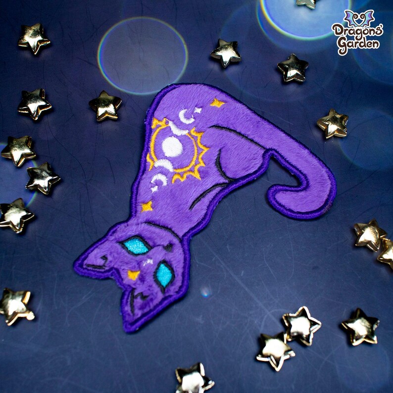 May include: A purple cat-shaped patch with a witch hat design. The hat features a sun, moon, and star motif in yellow and white. The cat's eyes are turquoise. The patch is surrounded by gold star-shaped embellishments. The text "Dragons Garden" is in the upper right corner.