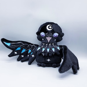 May include: A black plush crow with blue and white embroidered details, including a crescent moon on its head and stars on its wings.
