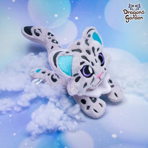 May include: A plush snow leopard toy with grey and white fur, blue eyes, and a white collar. The toy is lying on a white fluffy surface. The toy has a tag that says "Dragons' Garden".
