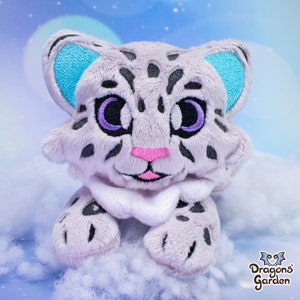 May include: A plush toy of a snow leopard with a white and grey fur pattern. The toy has blue ears, purple eyes, and a pink nose. The toy is sitting on a white fluffy surface.