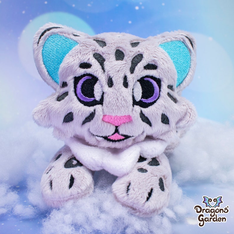 May include: A plush toy of a snow leopard with a white and grey fur pattern. The toy has blue ears, purple eyes, and a pink nose. The toy is sitting on a white fluffy surface.